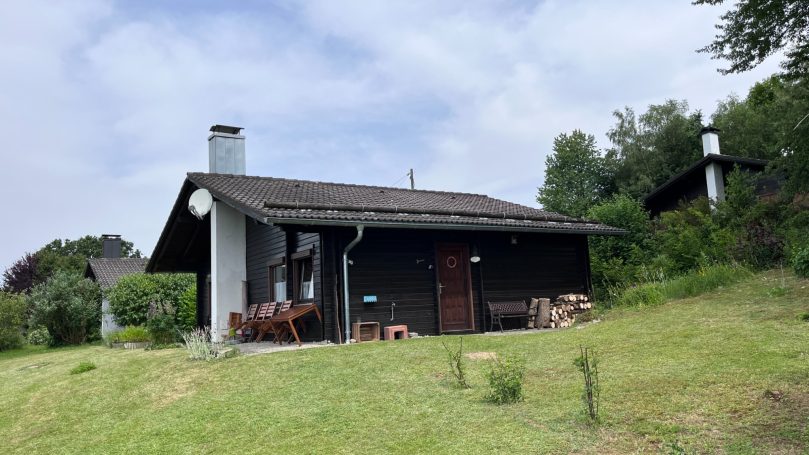Ein kleines, dunkles Holzhaus umgeben von grüner Landschaft unter bewölkem Himmel.