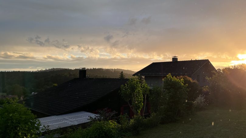 Sonnenuntergang über einem ländlichen Haus mit Bäumen im Vordergrund.