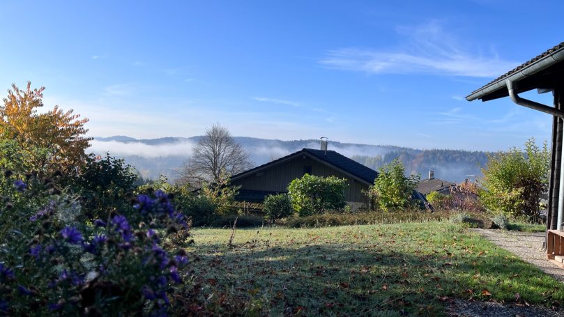 Landschaft mit einem Haus, grüner Wiese und Nebel in der Ferne bei klarem Himmel.
