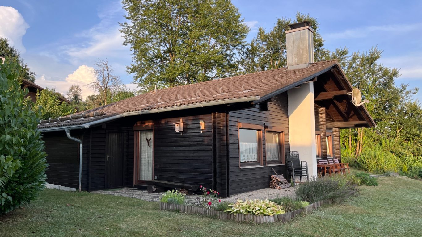 Ferienhaus Waldliebe Schwarzes Holzhaus mit Garten, umgeben von Bäumen und strahlend blauem Himmel.