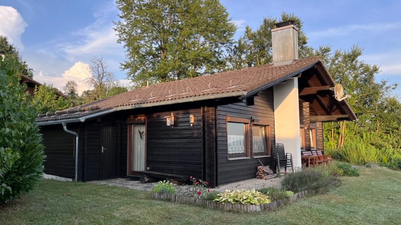 Schwarzes Holzhaus mit Garten, umgeben von Bäumen und strahlend blauem Himmel.