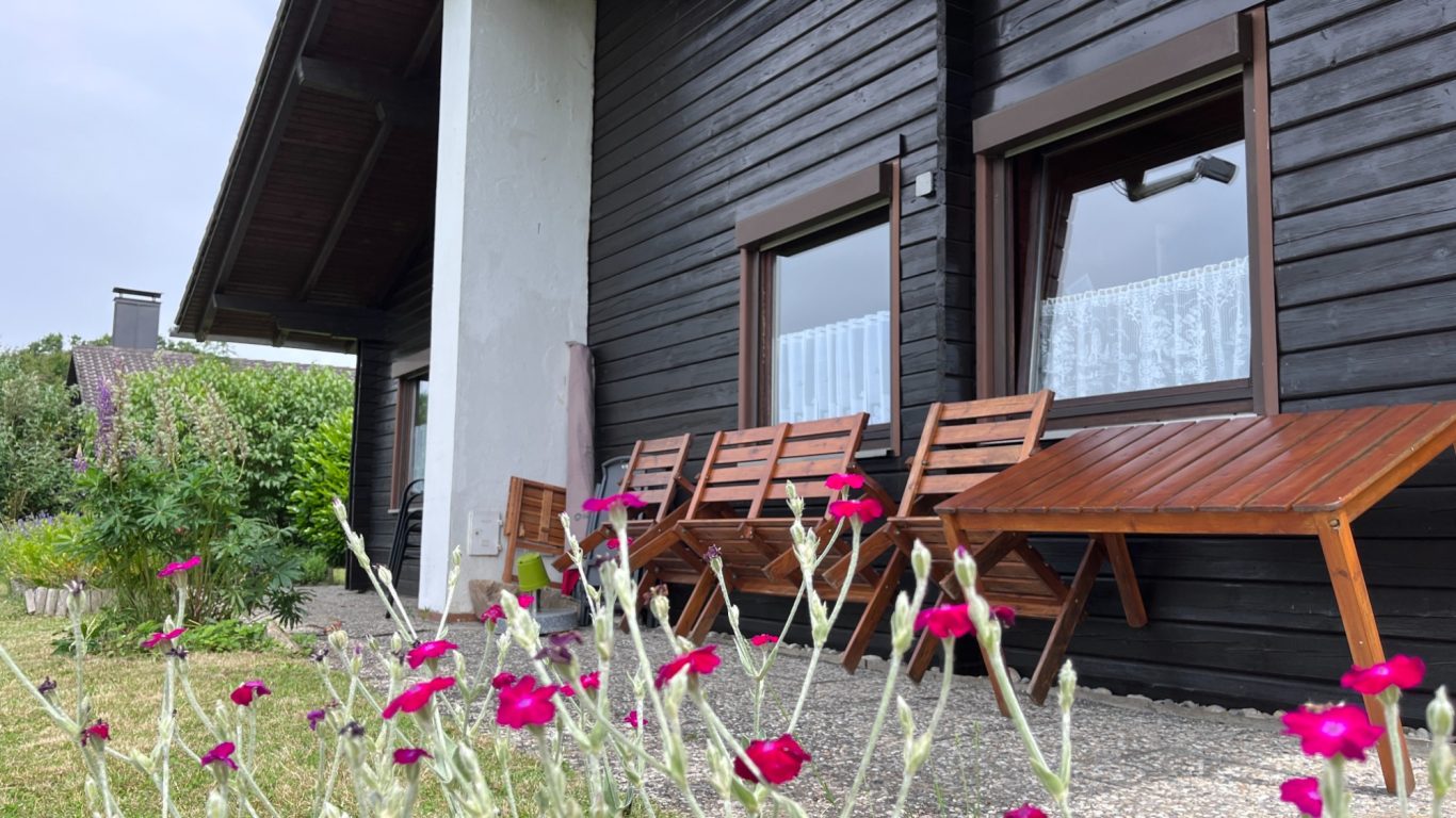 Alles wartet schon auf dich Zwei Holzliegen vor einem schwarzen Holzhaus, umgeben von bunten Blumen.