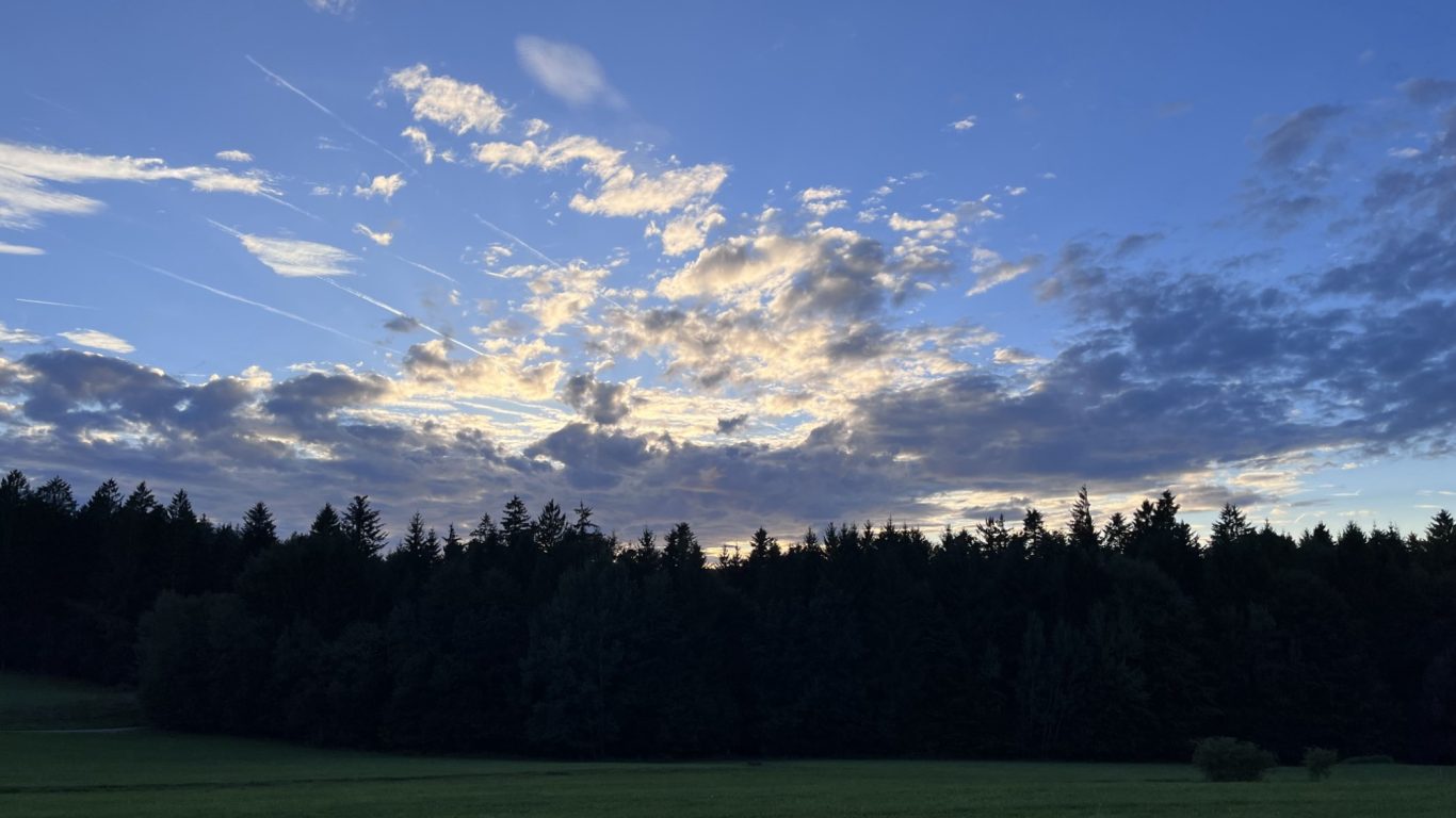 Abendhimmel mit Wolken über einem Wald, sanfte Lichtstrahlen durchbrechen die Dunkelheit.