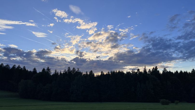 Abendhimmel mit Wolken über einem Wald, sanfte Lichtstrahlen durchbrechen die Dunkelheit.